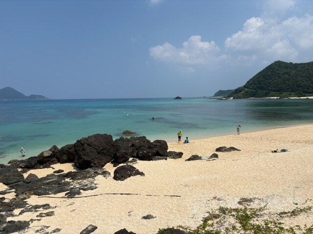 白い砂浜と黒い岩場が並ぶ海辺。浅瀬は淡いエメラルド色で、沖に向かうほど青が濃くなる。奥には緑に覆われた岬が突き出している